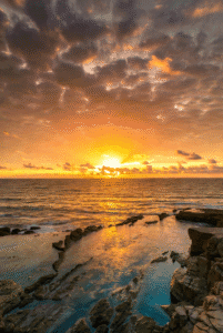Vibrant sunset over a turbulent ocean with orange light reflecting on dark, jagged rock formations in the foreground tide pool.