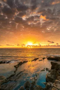 Vibrant sunset over a turbulent ocean with orange light reflecting on dark, jagged rock formations in the foreground tide pool.