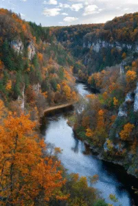 High-angle view of a dark, winding river cutting through a steep canyon with white limestone cliffs and dense forests displaying brilliant red and orange autumn foliage.