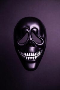 A glossy black mask with inverted teardrop-shaped eye holes and a wide, sinister grin revealing sharp white teeth. The mask’s reflective surface and dark purple backdrop create a dramatic and eerie visual, perfect for horror-themed art, Halloween costumes, or theatrical design.