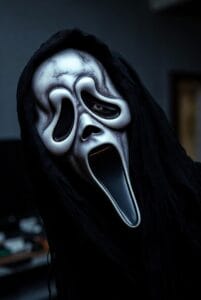 A close-up of a person wearing the iconic white Ghostface mask with hollow eyes, elongated nose, and a wide open mouth, framed by a black hood. The dimly lit, blurred background adds to the eerie and suspenseful atmosphere, evoking themes from the "Scream" horror movie franchise.